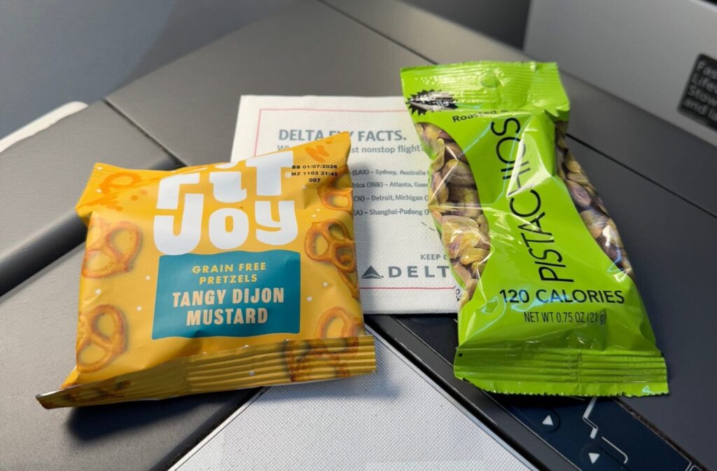 The image shows a tray table on an airplane with two snack packages and a card. One package is a yellow bag of "Utz Joy" grain-free pretzels with tangy Dijon mustard flavor. The other is a green bag of roasted and salted pistachios labeled as 120 calories. A Delta Air Lines card is partially visible behind the snacks.