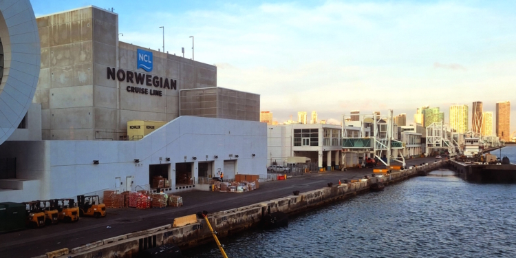 The image shows a waterfront scene with a large building labeled "Norwegian Cruise Line" on the left. The building is situated next to a dock area with several forklifts and stacks of goods. In the background, there are modern high-rise buildings under a clear blue sky. The water is visible in the foreground, indicating a port or harbor setting.