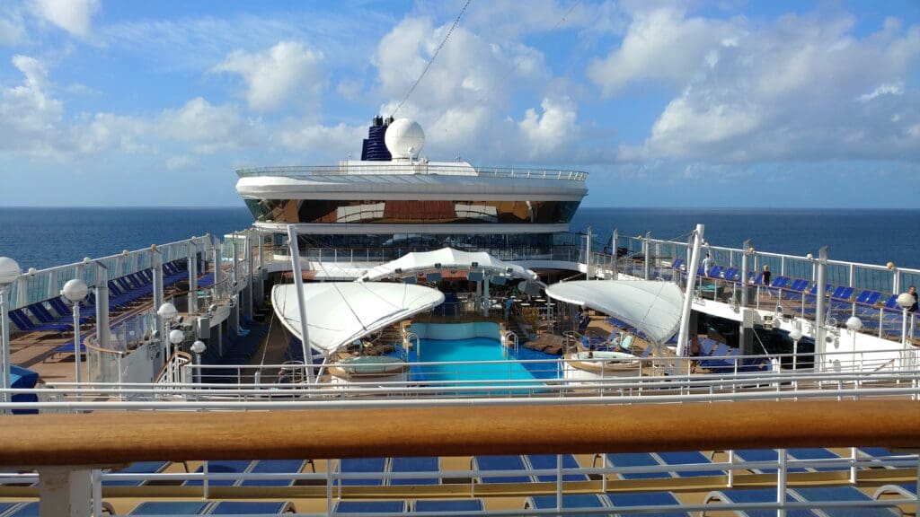 The image shows the deck of a cruise ship with a swimming pool in the center. There are lounge chairs arranged around the pool, and the ocean is visible in the background under a partly cloudy sky. The ship's upper structure and various amenities are also visible.