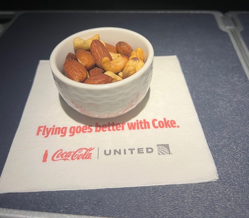 A small white bowl filled with mixed nuts, including almonds and cashews, is placed on a napkin. The napkin has red text that reads "Flying goes better with Coke." Below the text are the Coca-Cola and United Airlines logos. The setting appears to be an airplane tray table.