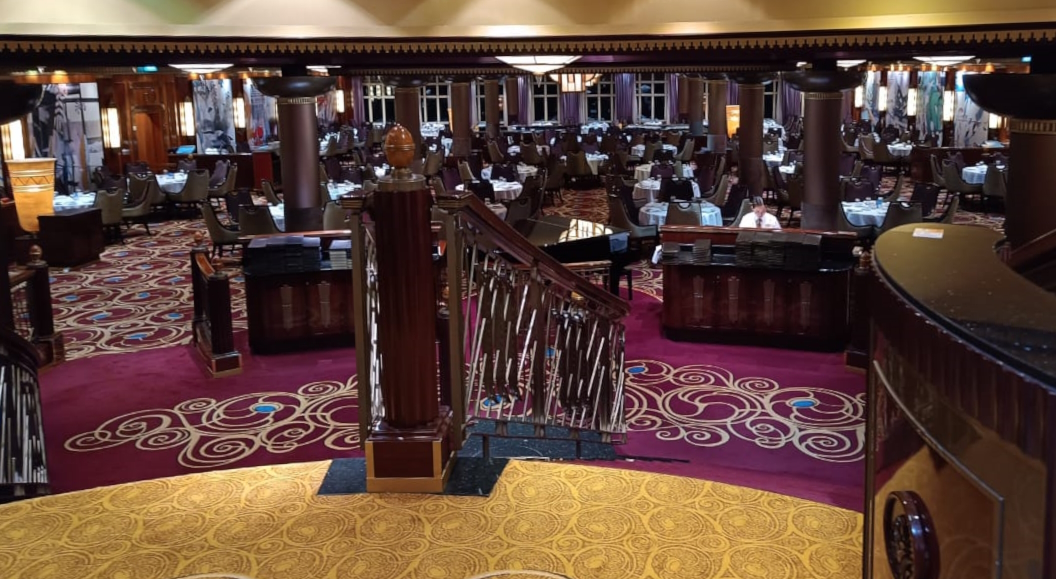 The image shows an elegant, spacious dining area with a richly decorated interior. There are numerous round tables set with white tablecloths and chairs arranged around them. The floor is covered with a patterned carpet in red and gold tones. A central staircase with ornate railings leads down into the dining area. The room is well-lit, with columns and decorative elements adding to the luxurious ambiance.