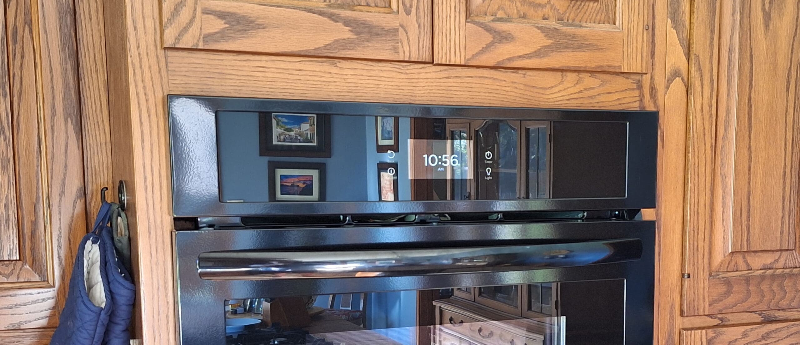 The image shows a modern black oven with a digital display showing the time as 10:56 AM. The oven is built into wooden cabinetry, and there are reflections of framed pictures and furniture on the oven's glass surface. A blue oven mitt is hanging on a hook to the left.