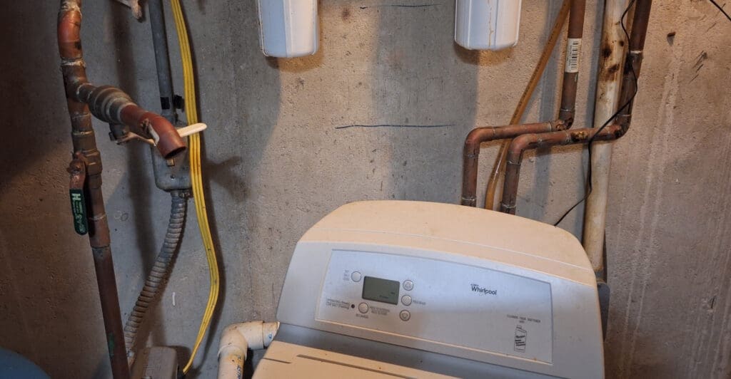 The image shows a basement or utility room setup with a water softener unit. The water softener is positioned against a concrete wall, with various pipes and electrical cables visible. The pipes are made of copper and are connected to the water softener. The unit has a control panel with buttons and a small display screen. Two white cylindrical components are mounted on the wall above the unit.