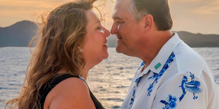 A couple is standing close together, smiling at each other with their noses touching. They are outdoors near a body of water, with a scenic sunset and mountains in the background. The woman has long, wavy hair and is wearing a sleeveless top, while the man is wearing a white shirt with blue floral designs.