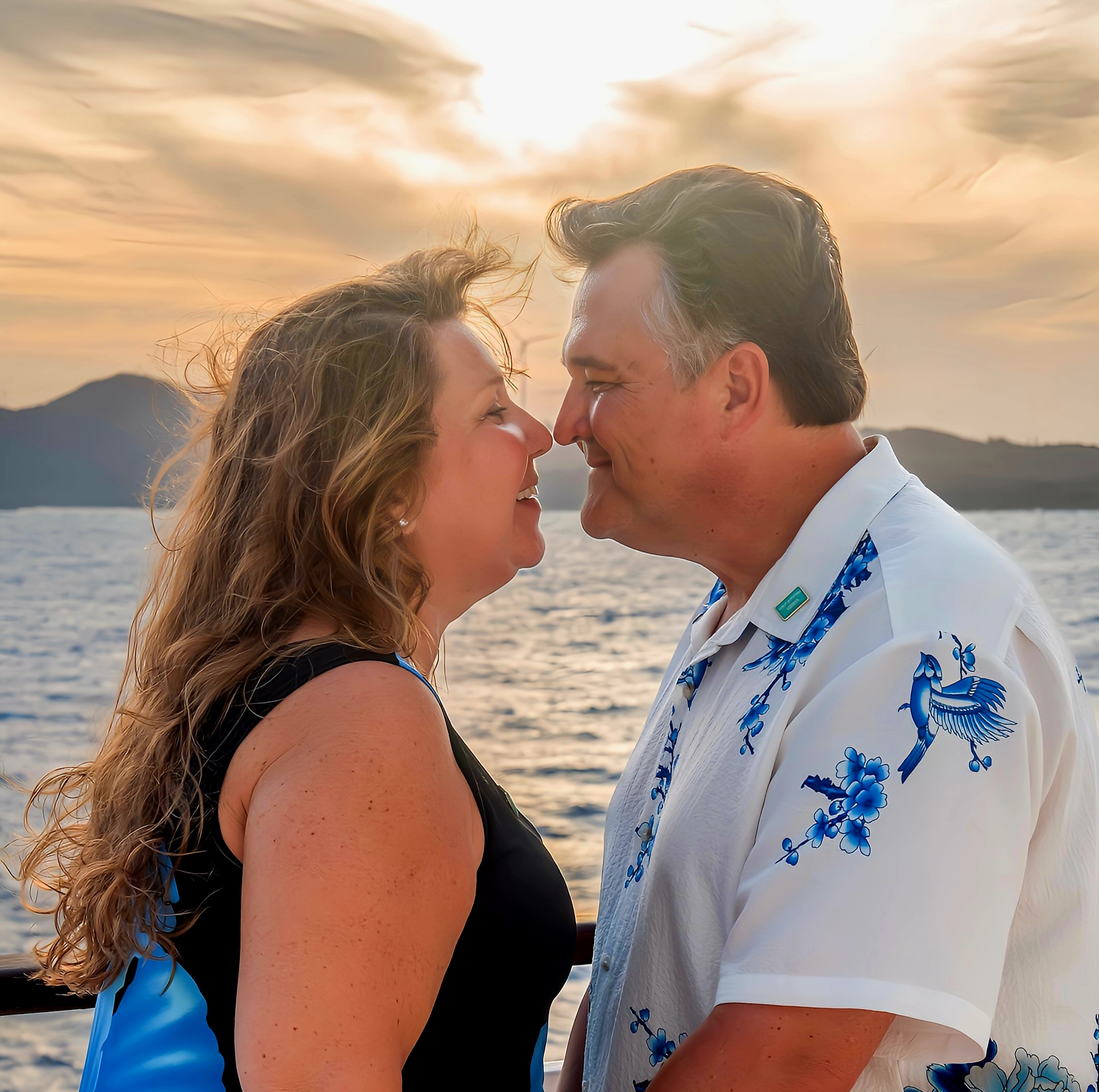 A couple is standing close together, smiling at each other with their noses touching. They are outdoors near a body of water, with a scenic sunset and mountains in the background. The woman has long, wavy hair and is wearing a sleeveless top, while the man is wearing a white shirt with blue floral designs.