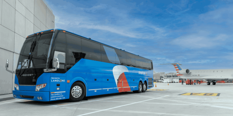 A blue bus with "American" branding is parked on an airport tarmac next to a building. The bus has "Operated by Landline" written on it. In the background, there is a white airplane with a red, white, and blue tail design. The sky is clear with some clouds.