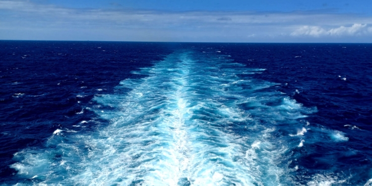 A vast ocean view with deep blue water and a trail of white foam left by a moving ship, extending towards the horizon under a partly cloudy sky.