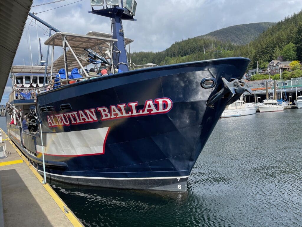 The image shows a large blue fishing boat named "Aleutian Ballad" docked at a marina. The boat has a prominent anchor at the front and is moored alongside a concrete pier. In the background, there are other boats and a hillside covered with trees and some buildings. The sky is partly cloudy.
