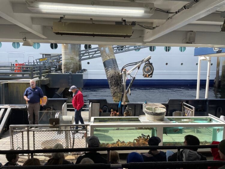 The image shows two men standing on a dock next to a large ship. One man is wearing a navy blue shirt and the other a red jacket. In front of them is a tank filled with crabs, and a group of people is seated in the foreground, watching the scene. The dock area has various equipment and a light fixture, and the ship in the background has circular windows.