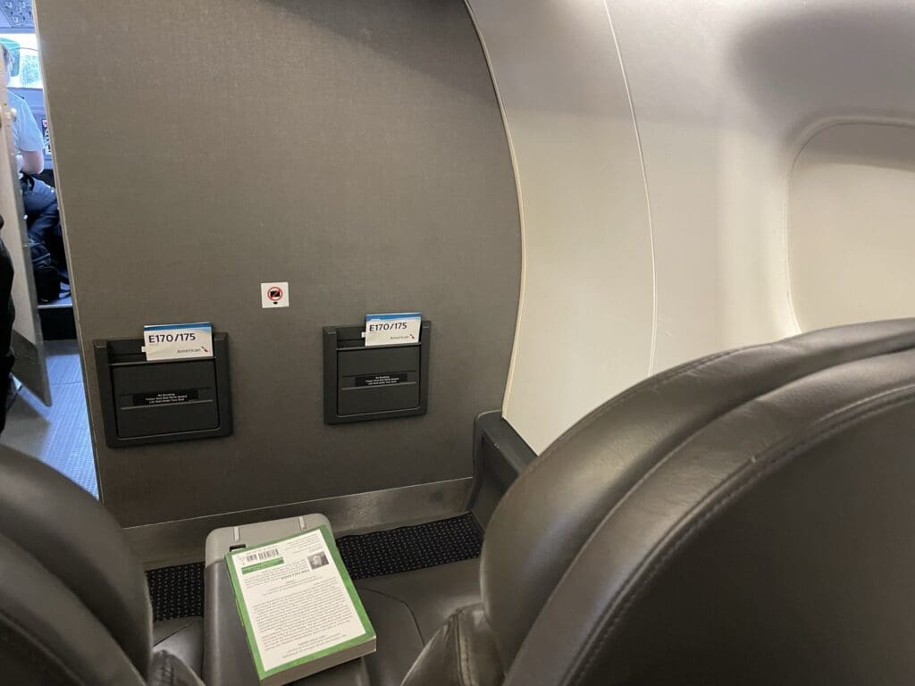The image shows the interior of an airplane, focusing on the back of a seat and the wall in front of it. There are two seatback pockets labeled "E170/175" with safety information cards. A book is placed on the armrest of the seat. Part of the cockpit and a pilot are visible through the open door on the left. The seats are upholstered in dark leather.