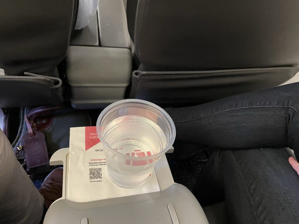 A plastic cup filled with water is placed on a tray table in an airplane. The cup is resting on a napkin with some text and a QR code. The image shows the back of an airplane seat and part of a person's legs wearing dark pants.