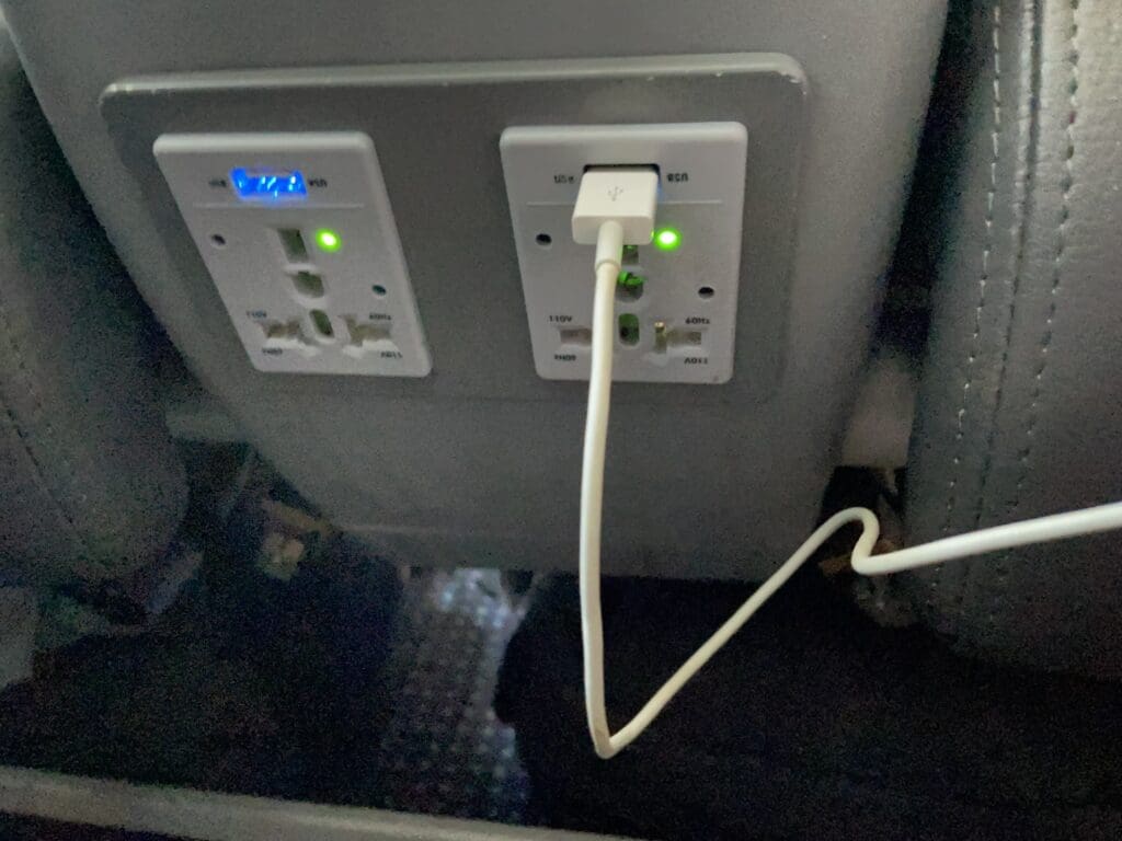 The image shows two electrical outlets on the back of an airplane seat. One outlet has a USB port with a cable plugged in, and both outlets have indicator lights. The surrounding area is part of the airplane's interior.