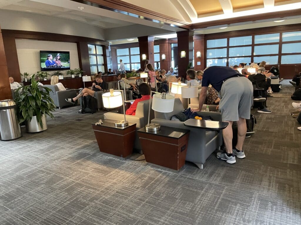 A spacious airport lounge with numerous people seated in comfortable chairs. Some individuals are using electronic devices, while others are conversing. The room features modern decor with wooden accents, potted plants, and soft lighting from table lamps. A television on the wall displays a program, and large windows allow natural light to enter the space.