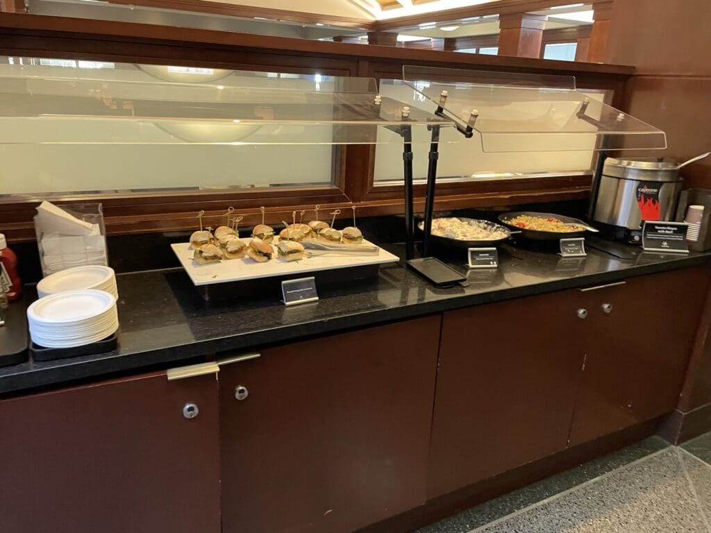 The image shows a buffet setup on a countertop. There are small sandwiches on a white tray with skewers, a bowl of pasta salad, and a bowl of another dish, possibly rice or noodles. There is a soup or beverage dispenser on the right. Plates, napkins, and condiments are on the left side of the counter. The area is enclosed with a glass or plastic sneeze guard.