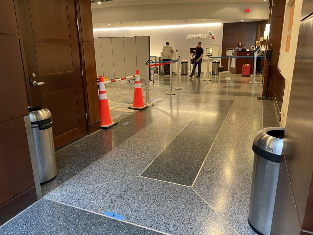 The image shows an airport check-in area with a counter in the background. There are people standing near the counter, and several orange traffic cones and a barrier tape are set up, possibly indicating a restricted area. The floor is made of polished stone, and there are trash bins on either side of the entrance. The word "American" is visible on the wall behind the counter.
