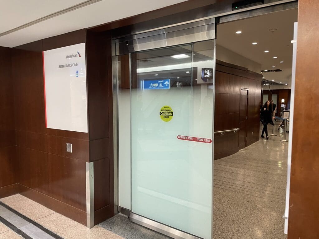 The image shows the entrance to an airport lounge, specifically the American Airlines Admirals Club. The entrance features a glass door with a yellow "Automatic Caution Door" sticker and a red "Automatic Door — Stand Clear" sign. The interior of the lounge is visible through the open door, with people walking inside. The walls are paneled with dark wood, and there is a sign with the Admirals Club logo on the left side of the entrance.
