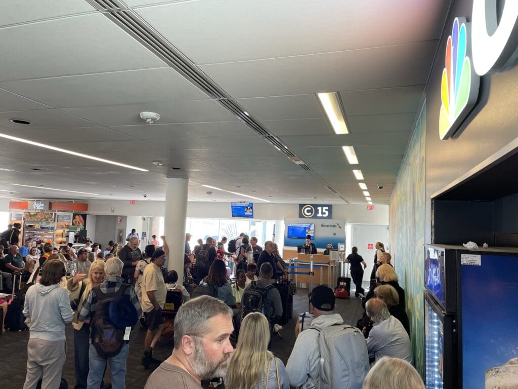 The image shows a busy airport gate area with many people waiting. There are passengers seated and standing, some with luggage. A gate sign displays "C 15" and an American Airlines logo. A vending machine is visible on the right, and a small shop or kiosk is in the background. The ceiling has modern lighting, and the atmosphere is bustling.