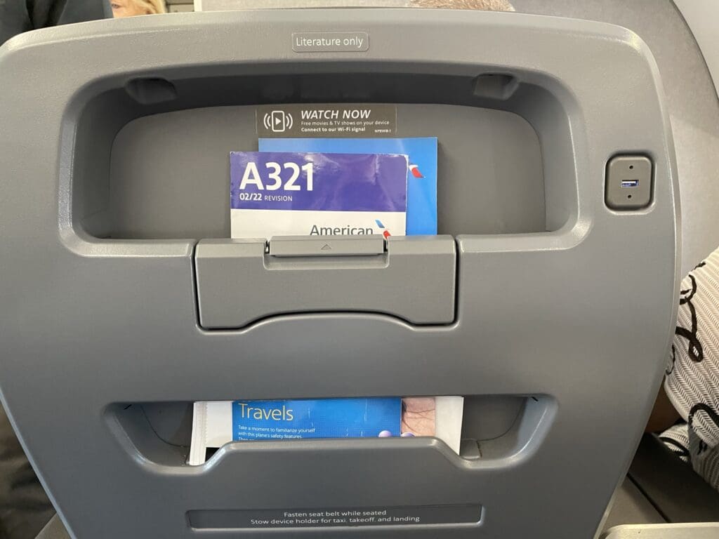 The image shows the back of an airplane seat with a literature pocket containing a safety information card labeled "A321 02/22 Revision" and a magazine titled "Travels." There is a USB port on the right side of the seatback, and a label at the top reads "Literature only." A small sign above the literature pocket mentions in-flight entertainment options.