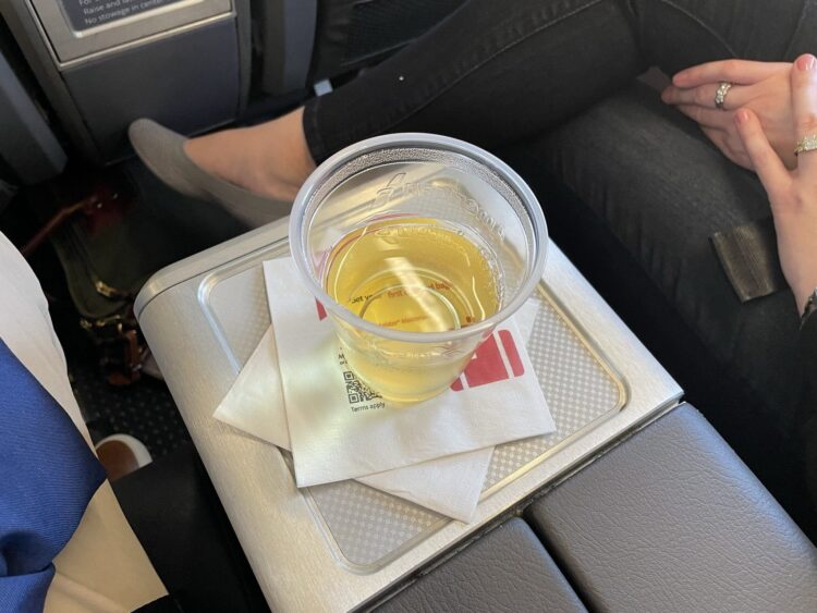 A plastic cup filled with a yellow beverage is placed on a tray table in an airplane. The cup is on top of a white napkin with a red logo. A person is seated next to the tray, with their hands resting on their lap, wearing a ring and a bracelet. The person is wearing dark pants and gray shoes.