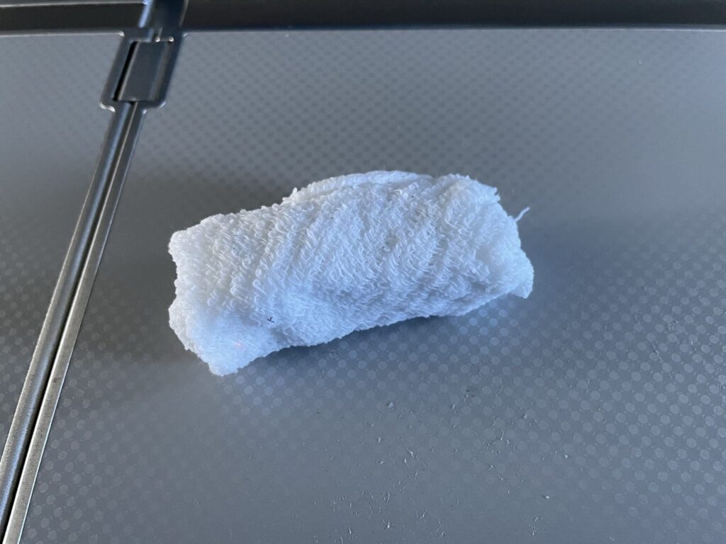 A small piece of white textured fabric is stuck to a gray, slightly reflective surface with a subtle dotted pattern. The fabric appears to be a bit crumpled and is positioned vertically.