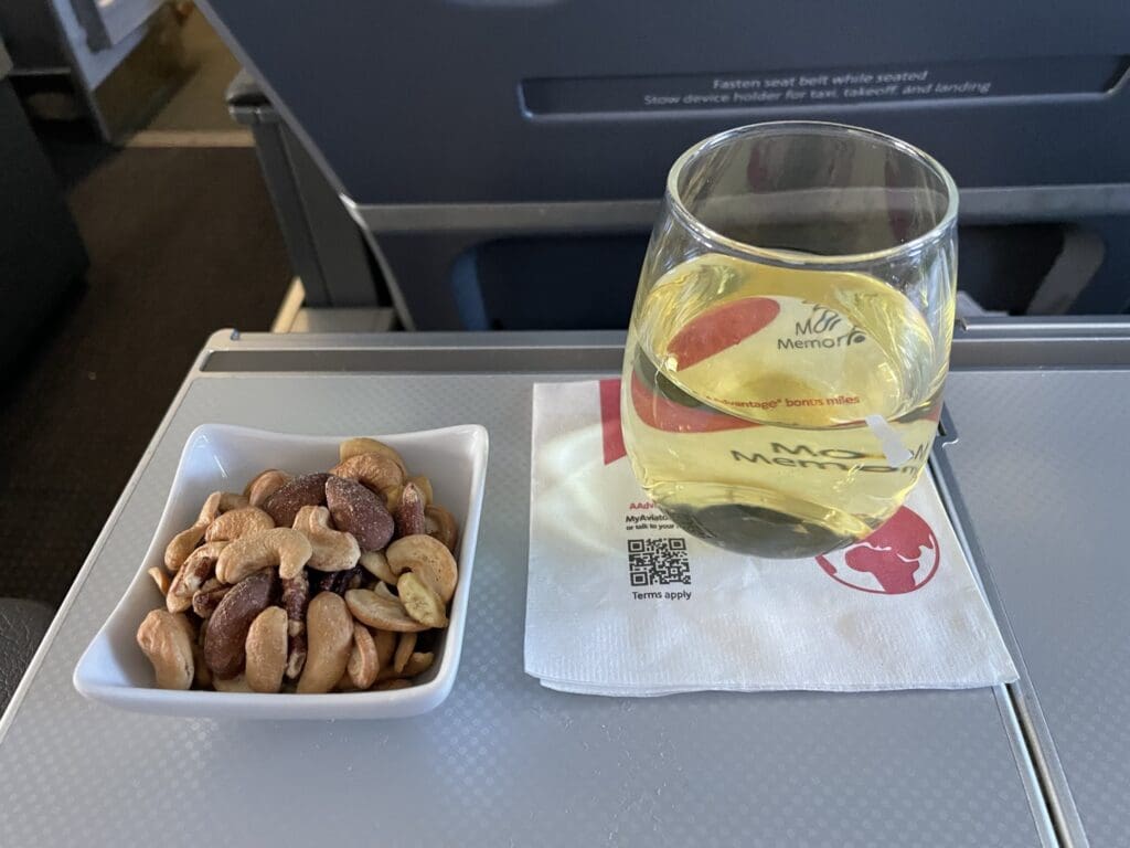 A glass of white wine and a small bowl of mixed nuts are placed on an airplane tray table. The glass is resting on a napkin with some text and a QR code. The background shows part of an airplane seat and the cabin interior.