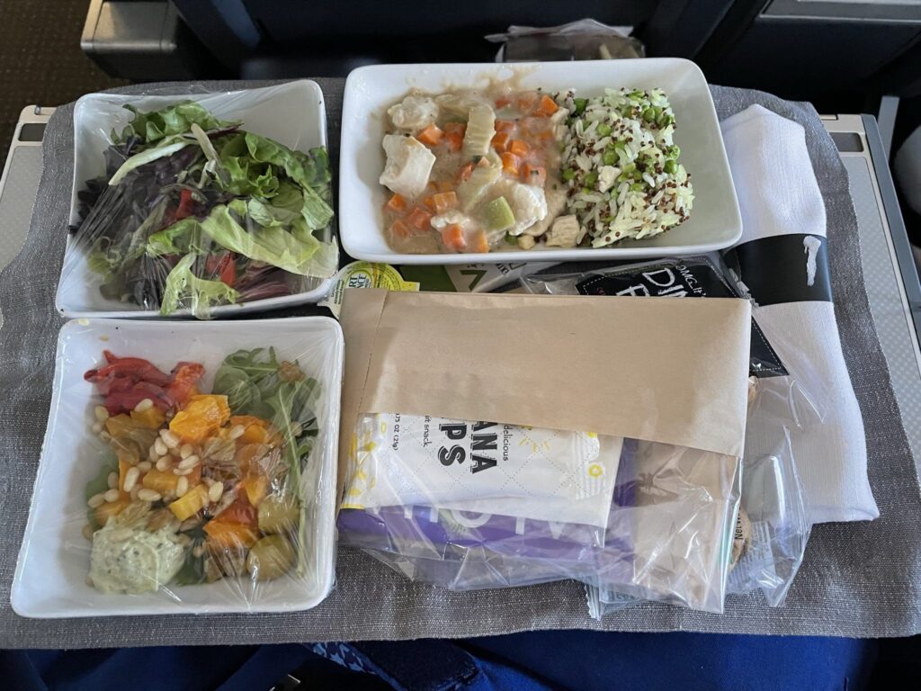 The image shows an airplane meal tray with various food items. There are three white dishes: one with a chicken and vegetable stew with rice, another with a mixed green salad, and a third with a salad containing colorful vegetables and beans, topped with a dollop of dressing. The dishes are covered with plastic wrap. Additionally, there are packaged items including banana chips, a napkin, and utensils. The tray is placed on a gray fabric mat.