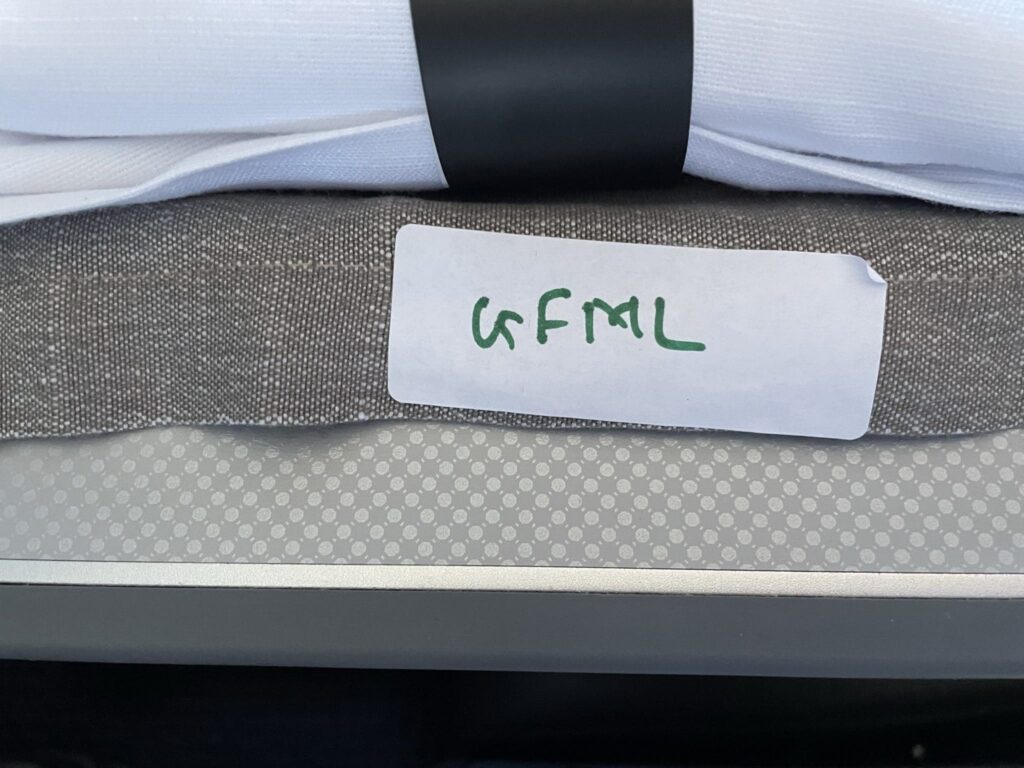 The image shows a close-up of a folded white napkin wrapped with a black band, placed next to a gray textured fabric. A white label with the letters "GFML" written in green is attached to the fabric. The background has a pattern of small dots.
