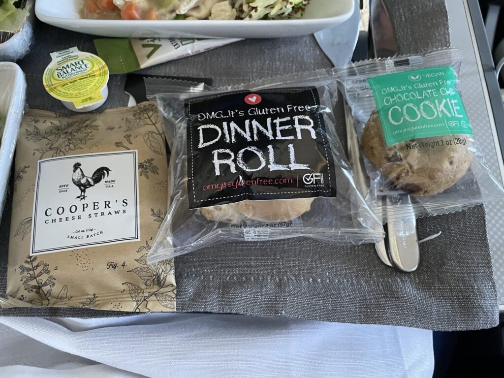 The image shows a meal setup on an airplane tray. There is a packaged gluten-free chocolate chip cookie and a gluten-free dinner roll. A packet of Cooper's cheese straws is also visible. A small container of Smart Balance spread is included. The items are placed on a gray fabric napkin with a metal knife and fork. Part of a salad or pasta dish is partially visible on the left.