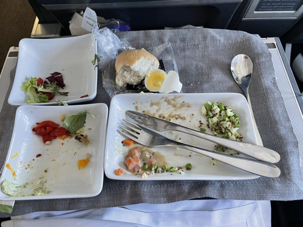The image shows a tray with three rectangular white plates containing remnants of a meal. The top plate has leftover peas, quinoa, and diced vegetables with a fork and knife resting on it. The bottom left plate has a few pieces of mixed salad leaves and red peppers. The bottom right plate has traces of sauce and a few pieces of red pepper. There is also a partially eaten bread roll with a small container of butter and a spoon on the tray, all placed on a gray fabric mat.