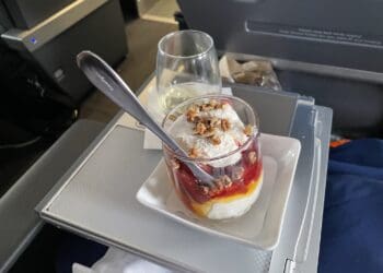 A dessert served on an airplane tray table. The dessert is in a glass cup, layered with whipped cream, fruit sauce, and topped with nuts. A spoon is placed in the cup. There is also a glass of white wine or champagne on the tray. The background shows part of the airplane interior.