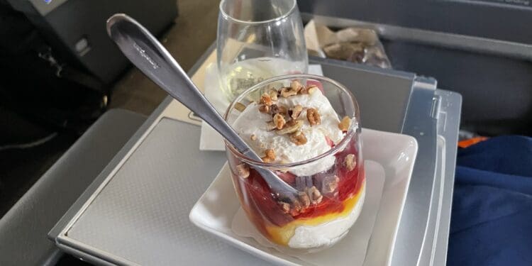 A dessert served on an airplane tray table. The dessert is in a glass cup, layered with whipped cream, fruit sauce, and topped with nuts. A spoon is placed in the cup. There is also a glass of white wine or champagne on the tray. The background shows part of the airplane interior.