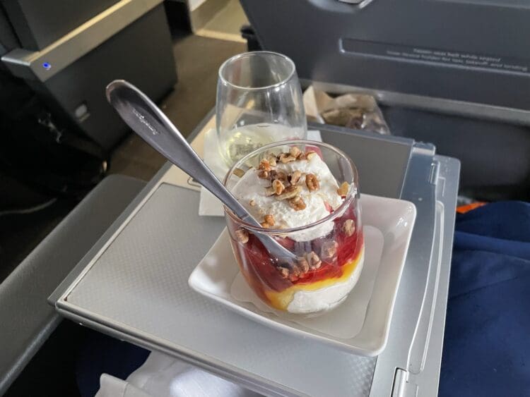 A dessert served on an airplane tray table. The dessert is in a glass cup, layered with whipped cream, fruit sauce, and topped with nuts. A spoon is placed in the cup. There is also a glass of white wine or champagne on the tray. The background shows part of the airplane interior.