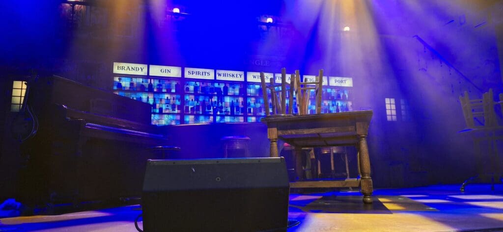 A dimly lit stage set with a bar theme. There is a piano on the left, a table with chairs stacked upside down on the right, and a microphone stand in the foreground. The background features shelves with bottles and signs labeled "Brandy," "Gin," "Spirits," "Whiskey," "Wines," "Cigars," and "Port." Blue and yellow stage lights create a moody atmosphere.