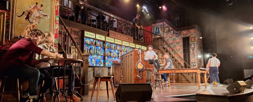 The image shows a lively stage set resembling a bar scene. There are several people interacting on stage, with some sitting at tables and others standing. The backdrop features a colorful bar with shelves of bottles and signs for different types of drinks like brandy, gin, and whiskey. A large, whimsical painting of a giraffe with a curly mustache is prominently displayed on the wall. The stage is well-lit with spotlights, and there are musical instruments, including a guitar, visible. The setting has a vibrant, theatrical atmosphere.