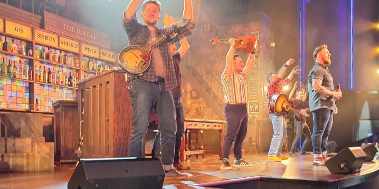 A lively stage performance is taking place with several musicians playing instruments like guitars and a banjo. They are energetically engaging with the audience, who are seated and clapping along. The stage is set up to resemble a bar, with shelves of bottles labeled "Brandy," "Gin," "Spirits," "Whiskey," "Wines," and "Fine Ales." The lighting is bright, creating a vibrant atmosphere.