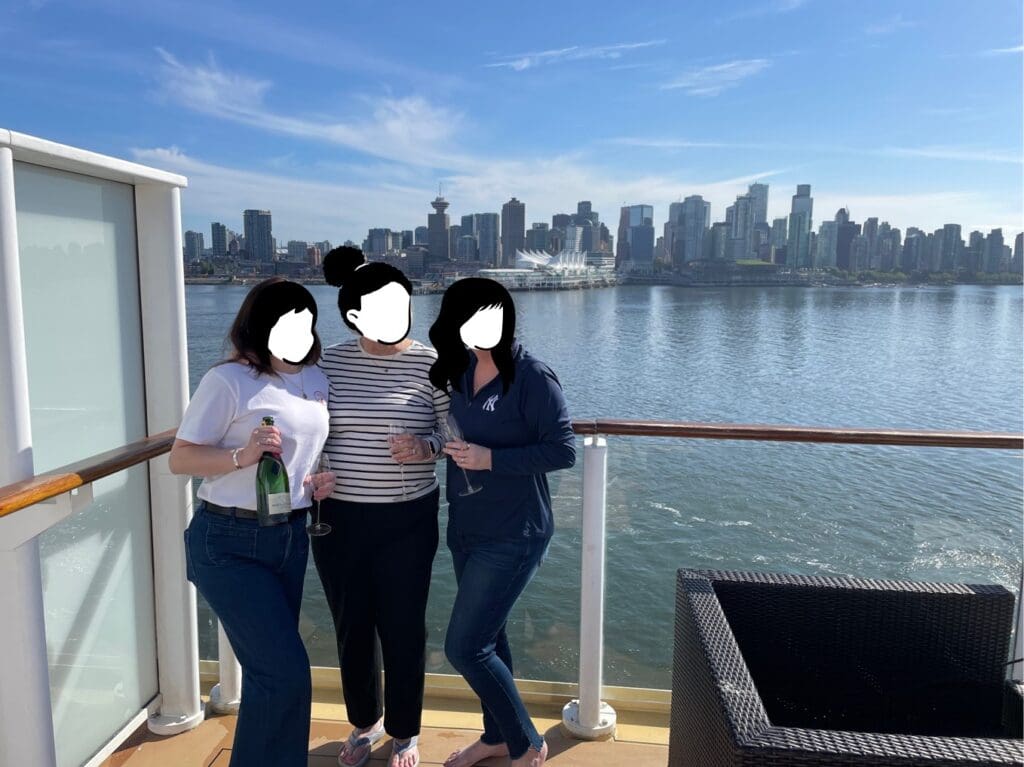 Three people are standing on a boat deck with a city skyline in the background. They are holding glasses and a bottle, possibly celebrating. The water and cityscape, including tall buildings, are visible under a clear blue sky.