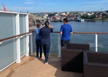 Three people are standing on the deck of a ship, looking out over a body of water. The deck has wooden flooring and a glass railing. In the background, there is an industrial area with cranes, shipping containers, and buildings along the waterfront. The sky is clear and blue.
