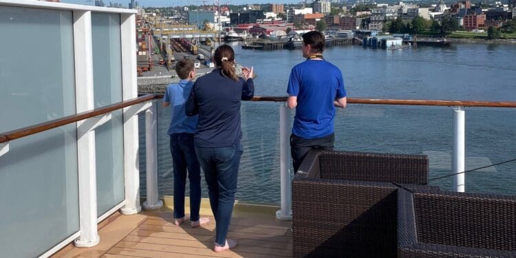 Three people are standing on the deck of a ship, looking out over a body of water. The deck has wooden flooring and a glass railing. In the background, there is an industrial area with cranes, shipping containers, and buildings along the waterfront. The sky is clear and blue.