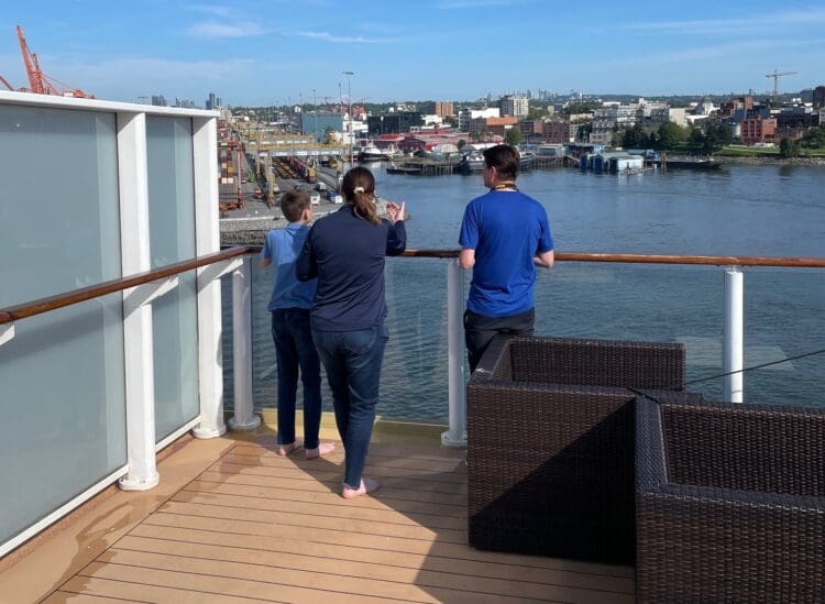 Three people are standing on the deck of a ship, looking out over a body of water. The deck has wooden flooring and a glass railing. In the background, there is an industrial area with cranes, shipping containers, and buildings along the waterfront. The sky is clear and blue.