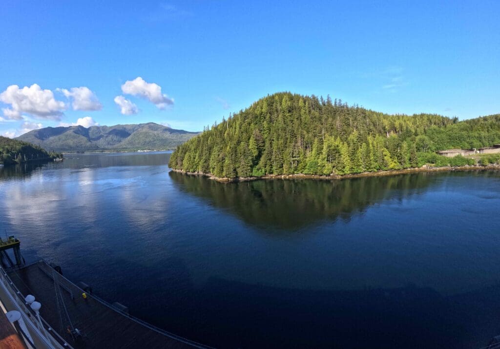 A scenic view of a calm body of water surrounded by lush, green forested hills under a clear blue sky with a few fluffy clouds. A dock or pier is visible in the foreground on the left side.