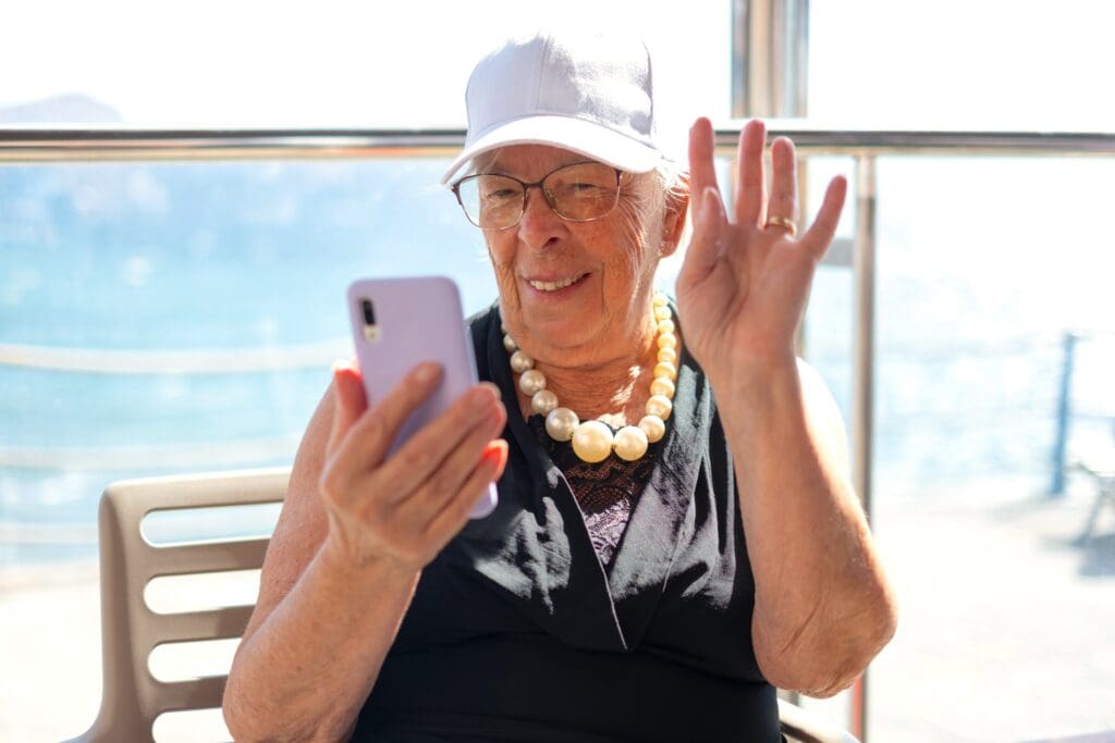 An elderly woman is sitting on a bench, smiling and waving at a smartphone she is holding, presumably having a FaceTime video call on a cruise ship. She is wearing a white cap, glasses, a black top, and a large pearl necklace. The background shows a bright, sunny day with a blurred view of water and railings.
