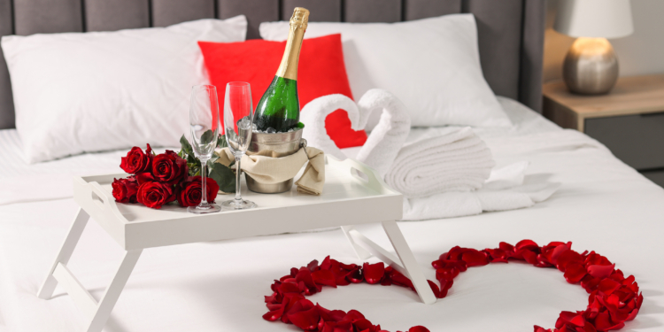 A romantic setup on a bed featuring a white tray with a bottle of champagne in an ice bucket, two empty champagne flutes, and a bouquet of red roses. The bed has white sheets, a red pillow, and towels folded into the shape of swans. Red rose petals are arranged in a heart shape on the bed. A bedside lamp is visible in the background.