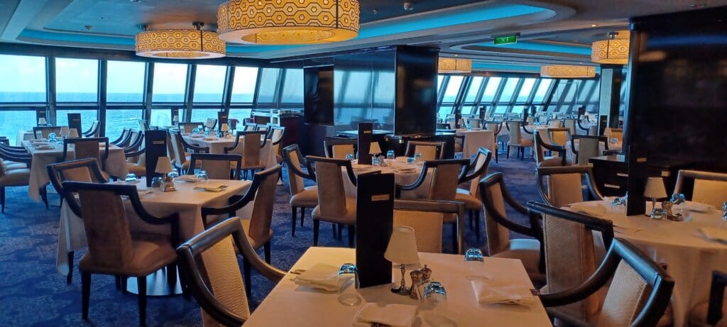 The image shows an elegant dining area with multiple tables set for a meal. Each table is covered with a white tablecloth and has neatly arranged place settings, including glasses, napkins, and small lamps. The room features large windows offering a view of the ocean, and the ceiling is adorned with decorative light fixtures. The chairs are upholstered and arranged around the tables, creating a spacious and inviting atmosphere.