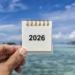 A hand is holding a small spiral-bound calendar displaying the year "2026" against a backdrop of a clear blue sky and a blurred view of the ocean.