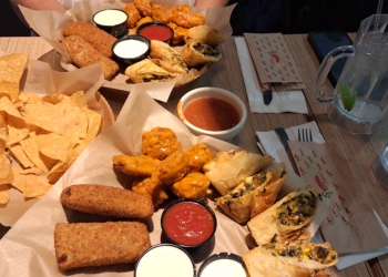 A table with a variety of appetizers, including tortilla chips, breaded sticks, buffalo wings, and stuffed spring rolls. There are several dipping sauces, including red and white ones. Two large mugs of water with lime slices are also on the table, along with napkins and utensils.
