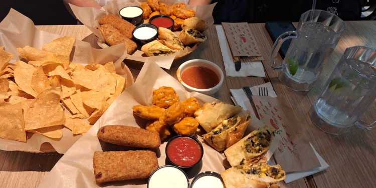 A table with a variety of appetizers, including tortilla chips, breaded sticks, buffalo wings, and stuffed spring rolls. There are several dipping sauces, including red and white ones. Two large mugs of water with lime slices are also on the table, along with napkins and utensils.
