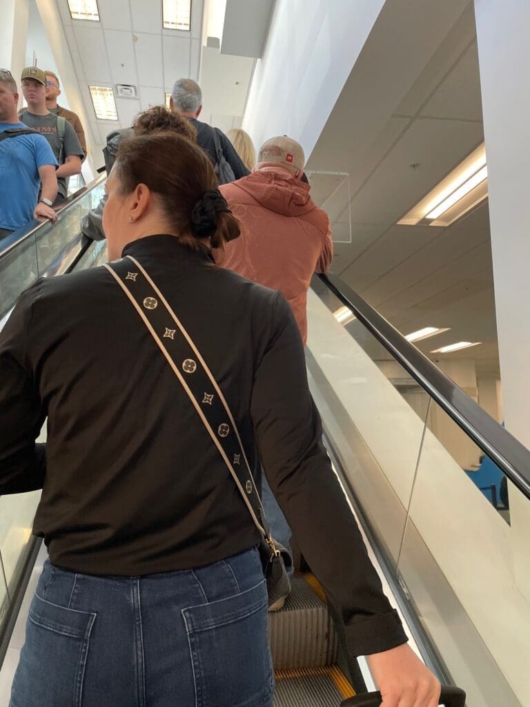 A group of people is standing on an escalator in a well-lit indoor setting. The person closest to the camera is wearing a black top and blue jeans, with a patterned strap across their back. Other individuals are visible ahead, some wearing casual clothing and hats. The ceiling has fluorescent lights.