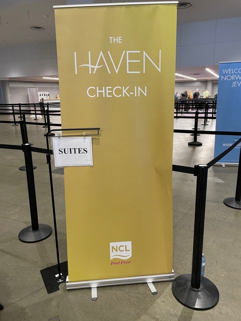 The image shows a yellow banner with the text "THE HAVEN CHECK-IN" and "NCL Feel Free" at the bottom. There is a sign attached to the banner that reads "SUITES." The banner is set up in an indoor area with stanchions and ropes, likely for organizing a queue. In the background, there is another blue banner with partially visible text and people standing in line.
