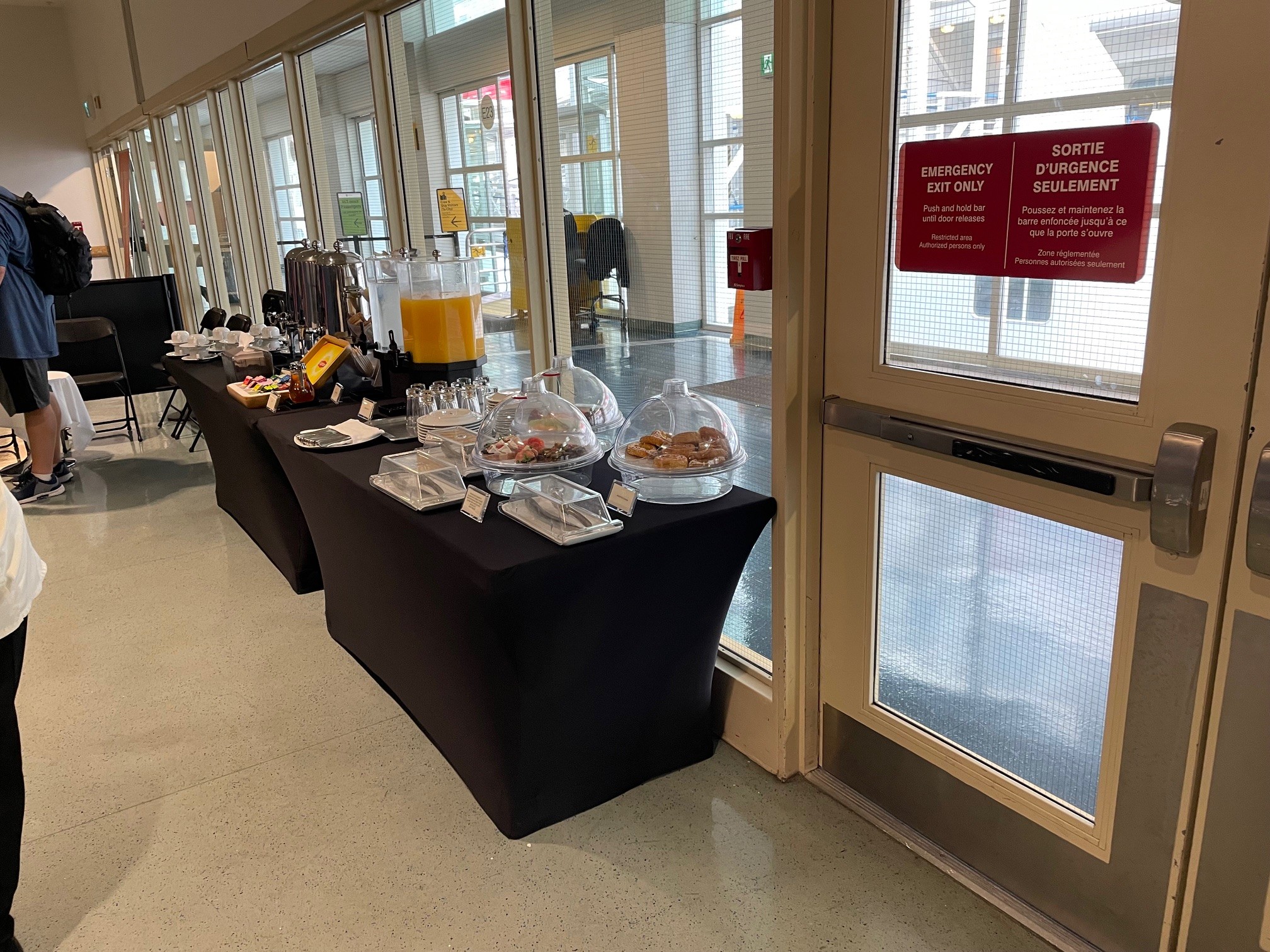 The image shows a buffet setup in a room with large windows. There are tables covered with black tablecloths, displaying various items for a breakfast or snack service. On the tables, there are dispensers with orange juice, coffee urns, cups, plates, and covered trays with pastries and other food items. There are also small signs or labels in front of the items. To the right, there is an emergency exit door with red signs indicating it is for authorized personnel only.