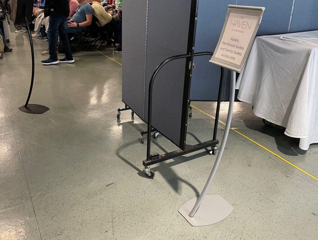 The image shows a room with many people seated in rows of chairs, suggesting a meeting or event. A man is walking down the aisle. On the right, there is a partition and a sign on a stand that reads "The Haven by Norwegian, Haven, Penthouse Suites and Family Suites access only." A table with a white tablecloth is partially visible behind the partition.
